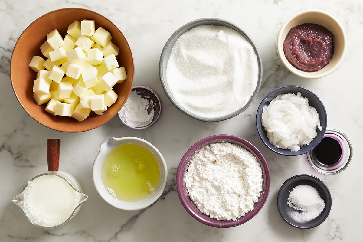 Overhead shot of neatly arranged baking ingredients, cubed butter, sugar, eggs, flour, oil, ube extract, and other components set in small bowls on a marble countertop, ready for making the cake batter.