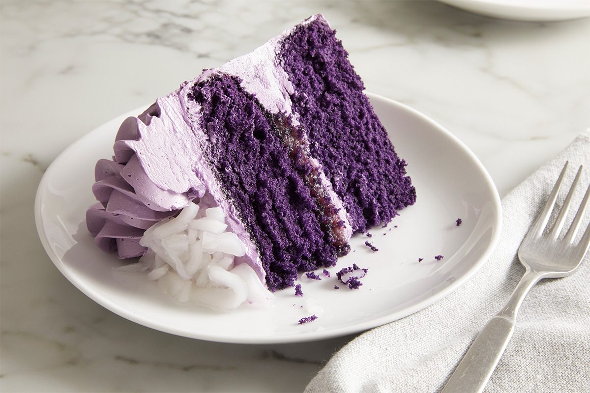 Close up shot of a plated slice of Ube Cake showcasing its bold purple crumb and layers of frosting, with a fork and linen napkin arranged beside the plate