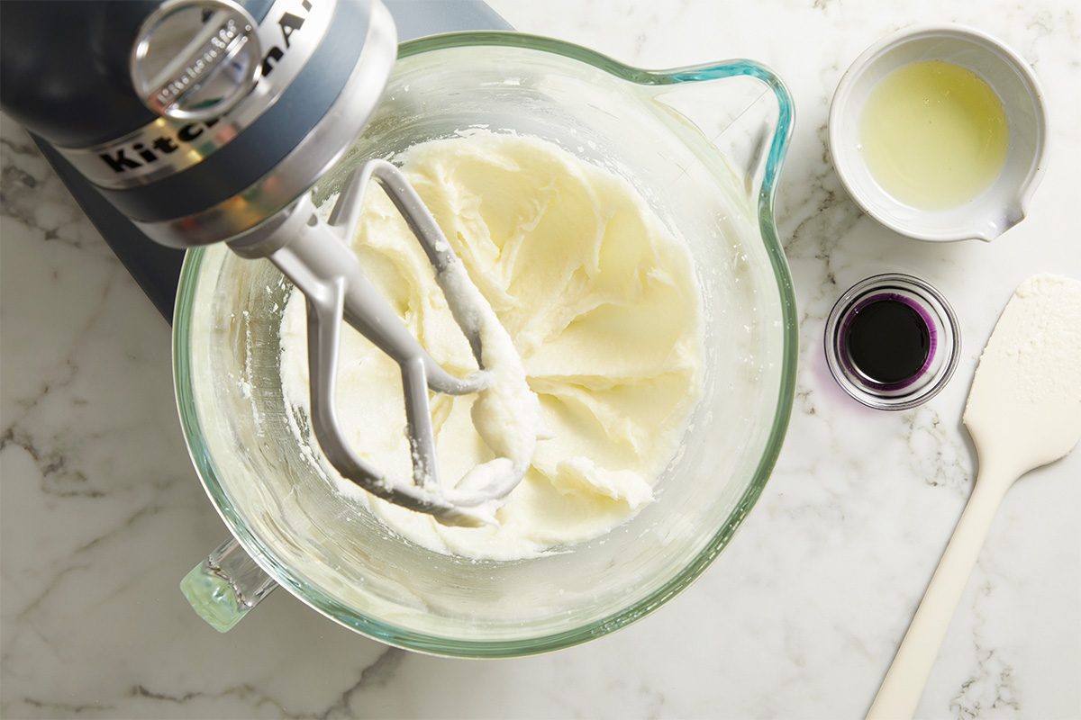 Overhead shot of a stand mixer bowl filled with softened butter being whipped to a pale, creamy texture, with vanilla and other liquids nearby in small measuring cups.