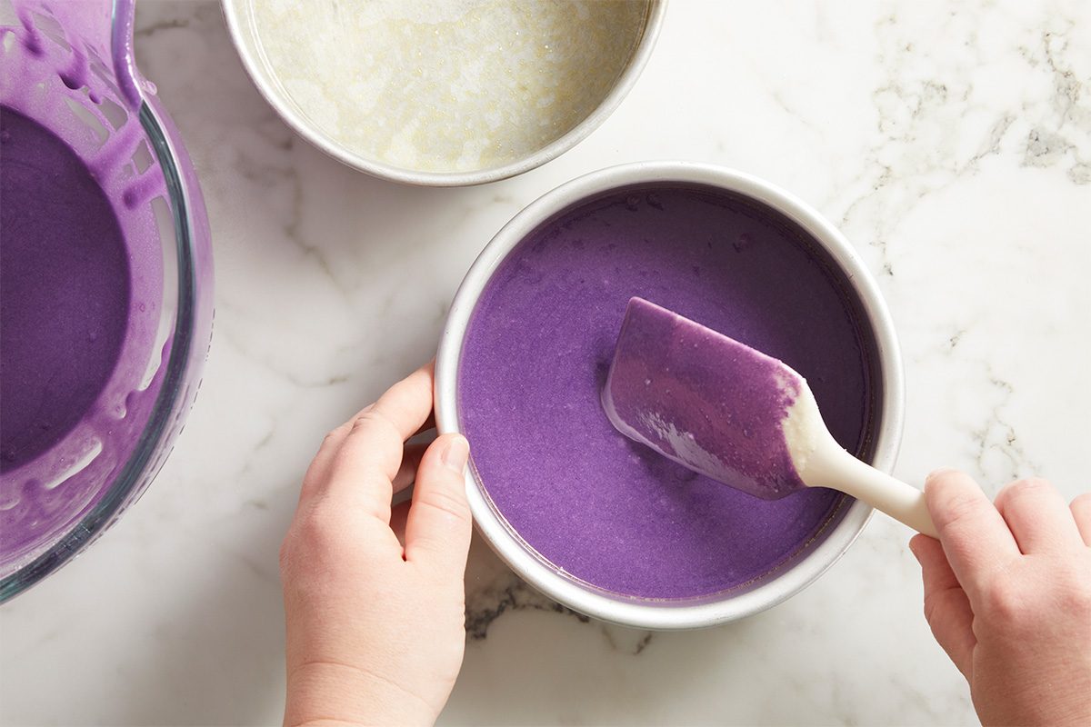 Overhead shot of a pair of hands spreading thick purple cake batter evenly into a round cake pan using a silicone spatula.