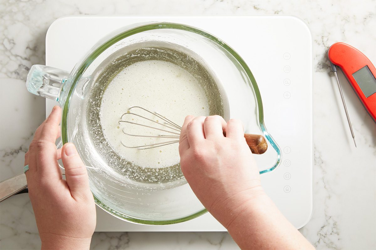 Overhead shot of dry ingredients being sifted into a mixing bowl, with hands holding the sifter and whisk surrounded by baking tools on a white cutting board.