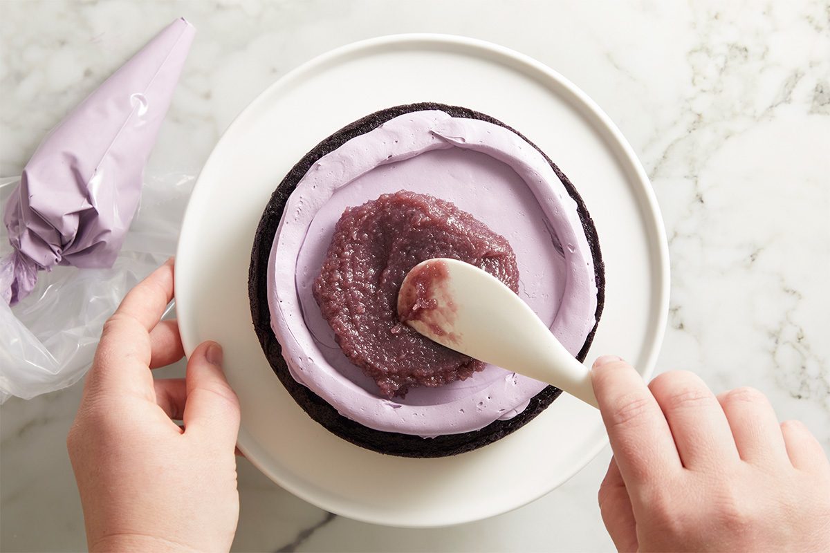 Overhead shot of hands spreading a layer of purple frosting over a chilled cake layer on a white cake plate, creating a smooth and even surface.