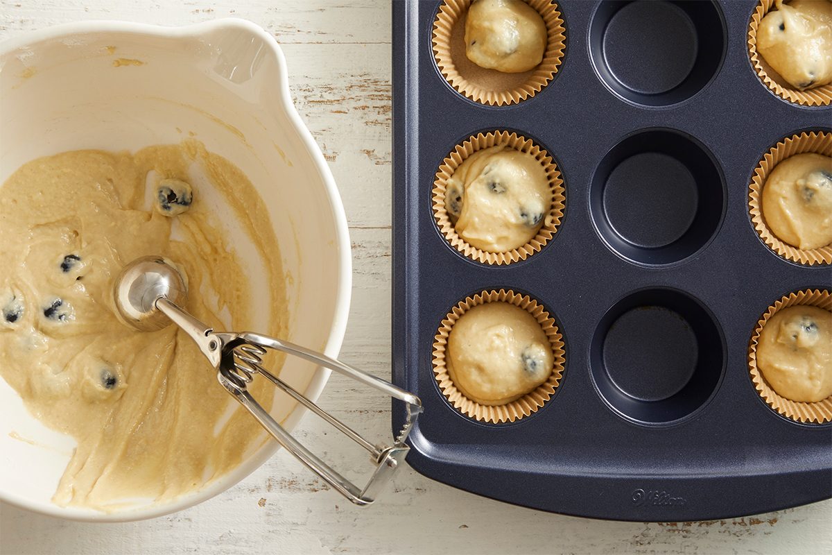 A mixing bowl with muffin batter and a scoop sits beside a muffin tin partially filled with paper liners and blueberry muffin batter.