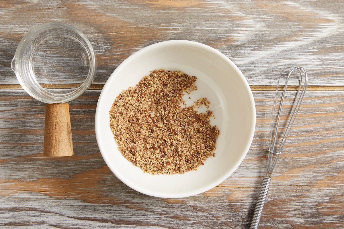 Overhead shot of a bowl of ground flaxseed sits next to a metal whisk and a small glass cup with a wooden handle on a rustic wooden board.