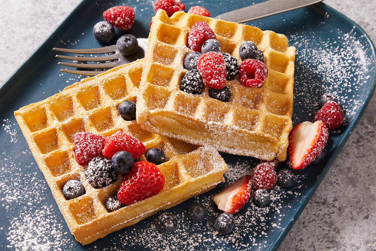 Close-up shot of Vegan Waffles show two square waffles topped with powdered sugar strawberries blueberries and raspberries on a dark blue plate beside a fork and knife extra berries are scattered around the plate
