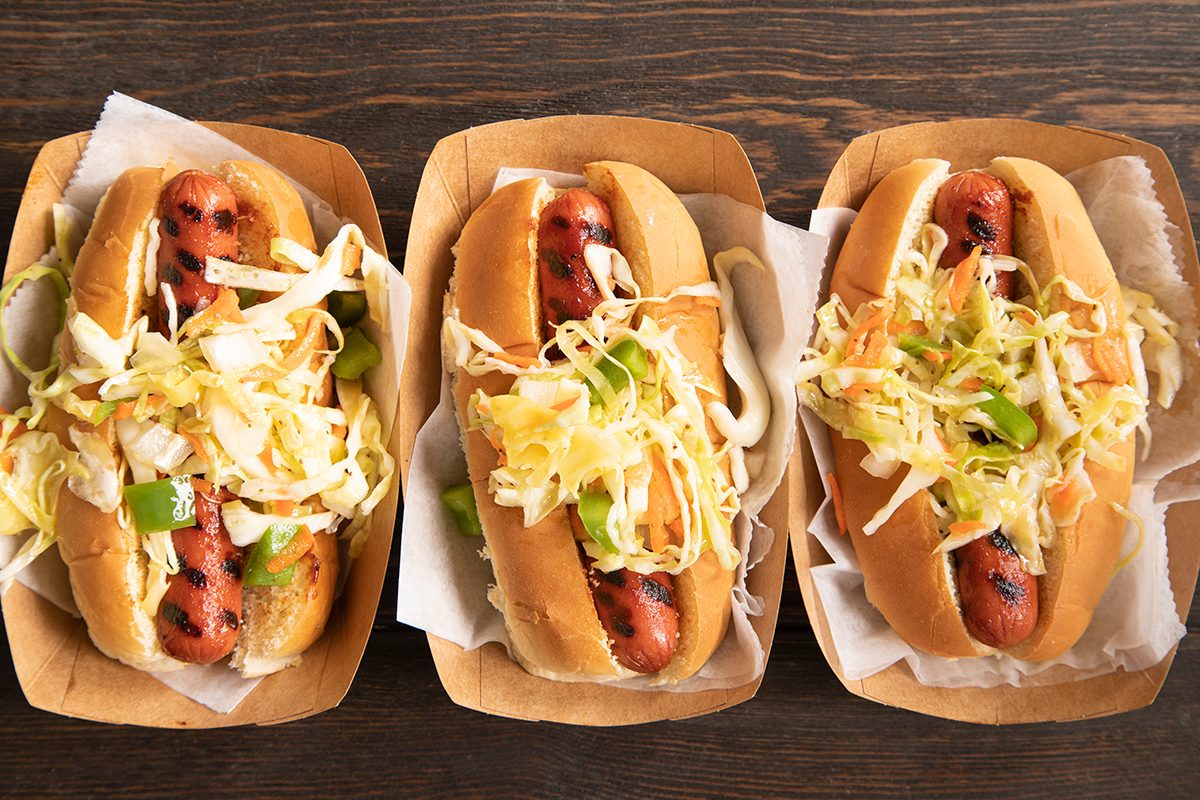 Three grilled hot dogs in buns are topped with shredded lettuce and diced vegetables, each served in a brown paper tray lined with parchment paper on a wooden surface.