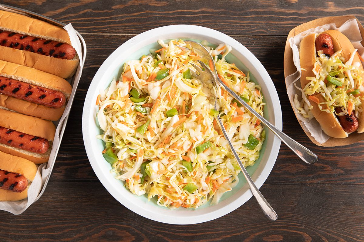A bowl of coleslaw with two serving forks sits on a wooden table, flanked by a tray of grilled hot dogs in buns on the left and two assembled hot dogs with coleslaw on the right.