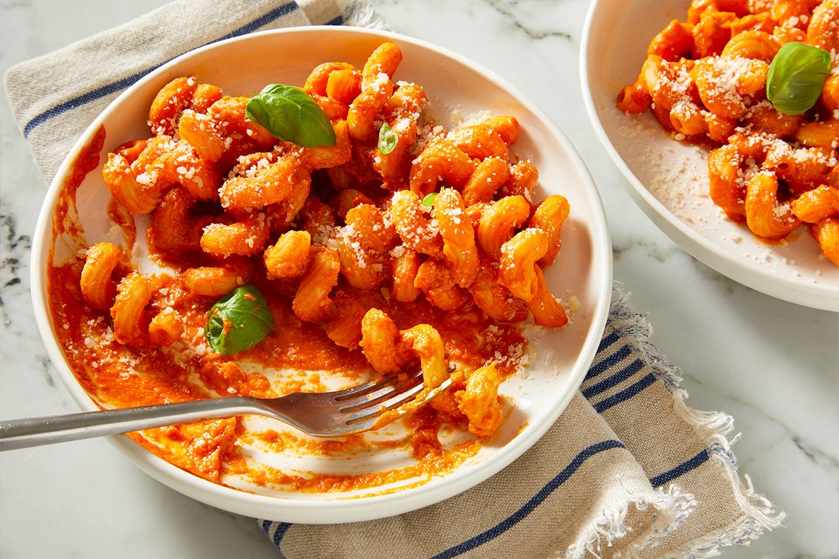 Overhead close-up of vodka pasta being served from a bowl with a spoon, highlighting the creamy tomato sauce texture and short-cut pasta shape. Finished with grated Parmesan and fresh herbs.