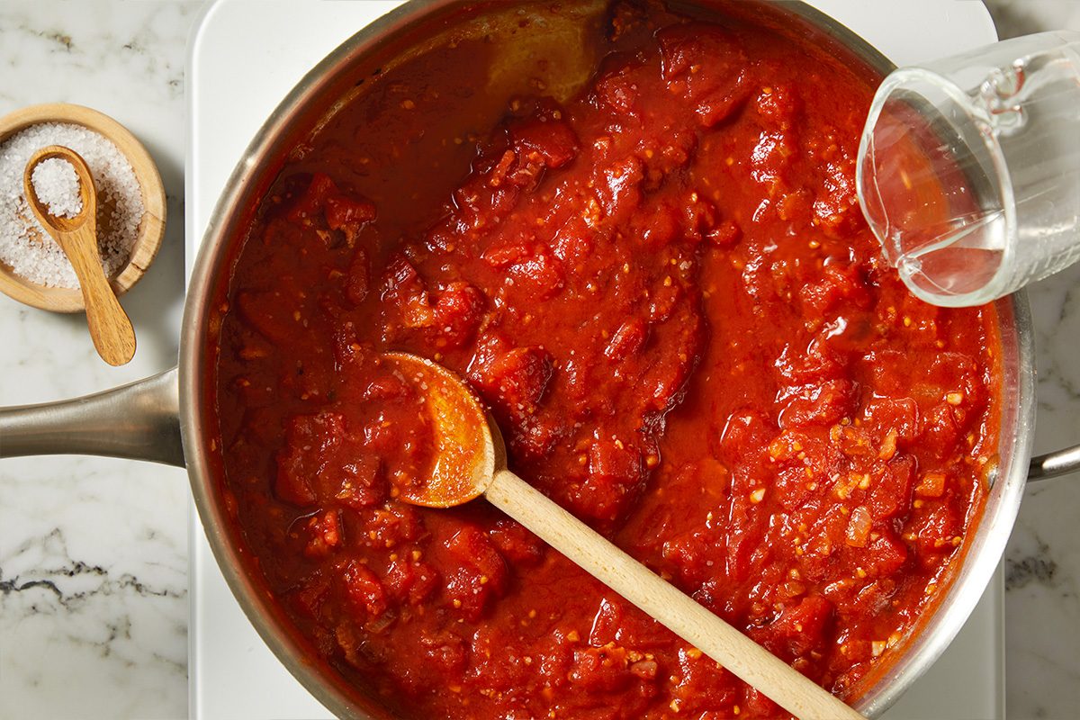 Overhead shot of crushed whole plum tomatoes added to the skillet, forming a vibrant red sauce as vodka is poured in. The tomato mixture simmers with bacon and aromatics, beginning to transform into a classic vodka sauce.
