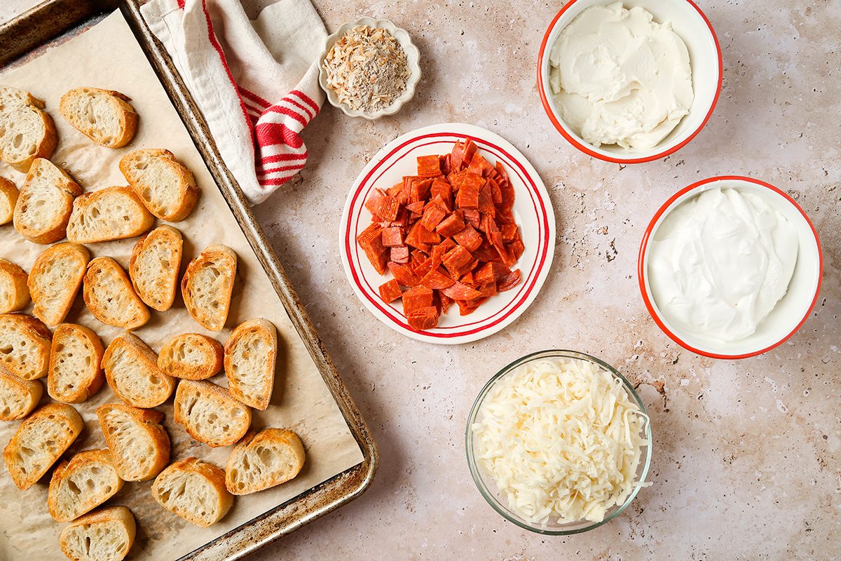 A baking tray with toasted baguette slices sits on a counter next to bowls containing diced pepperoni, shredded cheese, cream cheese, and sour cream. A red-striped towel and a small bowl of dried herbs are also visible.