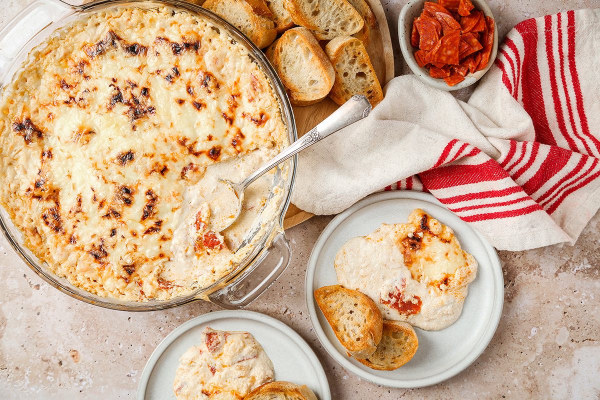 A dish of baked creamy dip with a spoon, surrounded by sliced baguette, a bowl of sun-dried tomatoes, and two plates with bread and dip, on a light surface with a red-striped napkin.