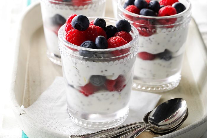 Three clear glasses filled with layers of creamy yogurt, chia seeds, and fresh blueberries and raspberries, placed on a tray next to three metal spoons.