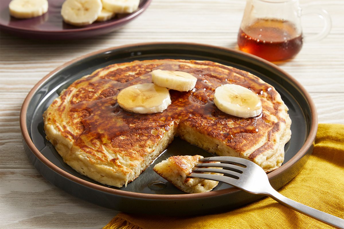 Overhead shot of a stack of Banana pancakes topped with banana slices, with a wedge cut out and placed in the foreground. A fork rests beside the plate, and a syrup jar and banana slices are visible in the background.