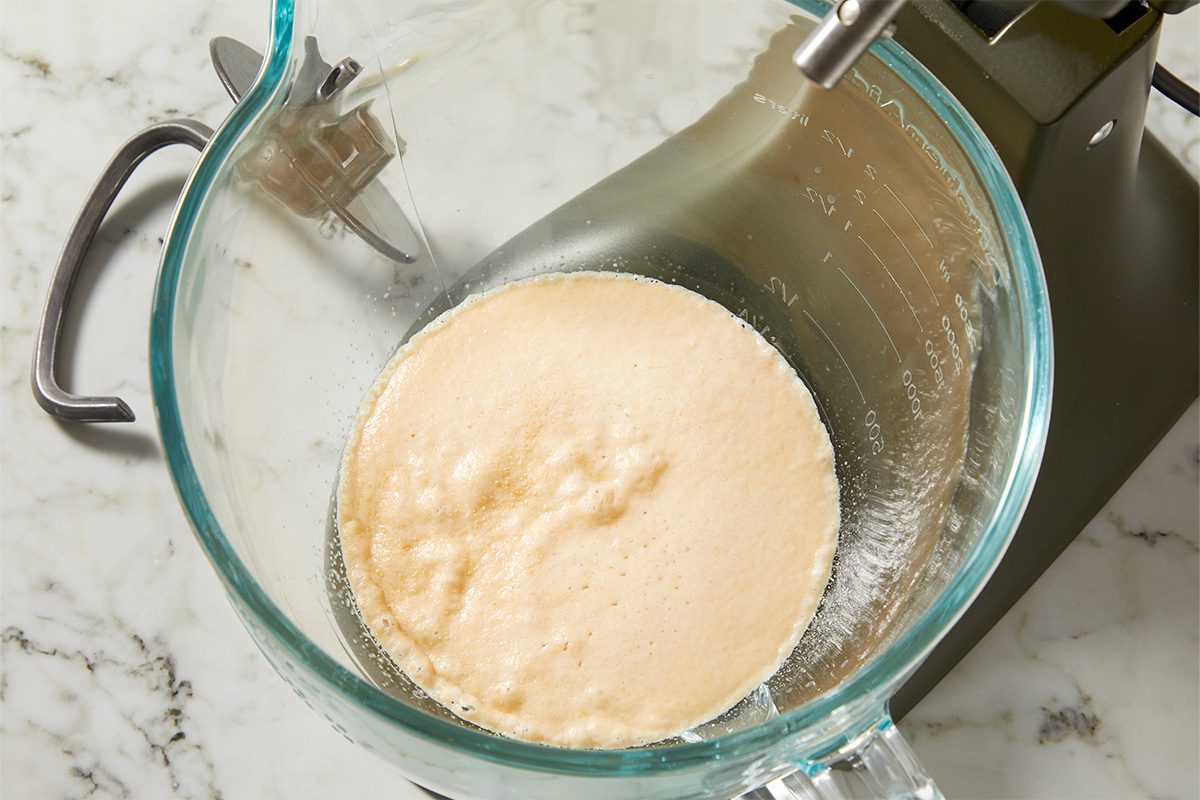 Overhead view of warm milk in a glass mixing bowl, the first step in preparing carrot cake cinnamon roll dough, photographed on a light marble surface.