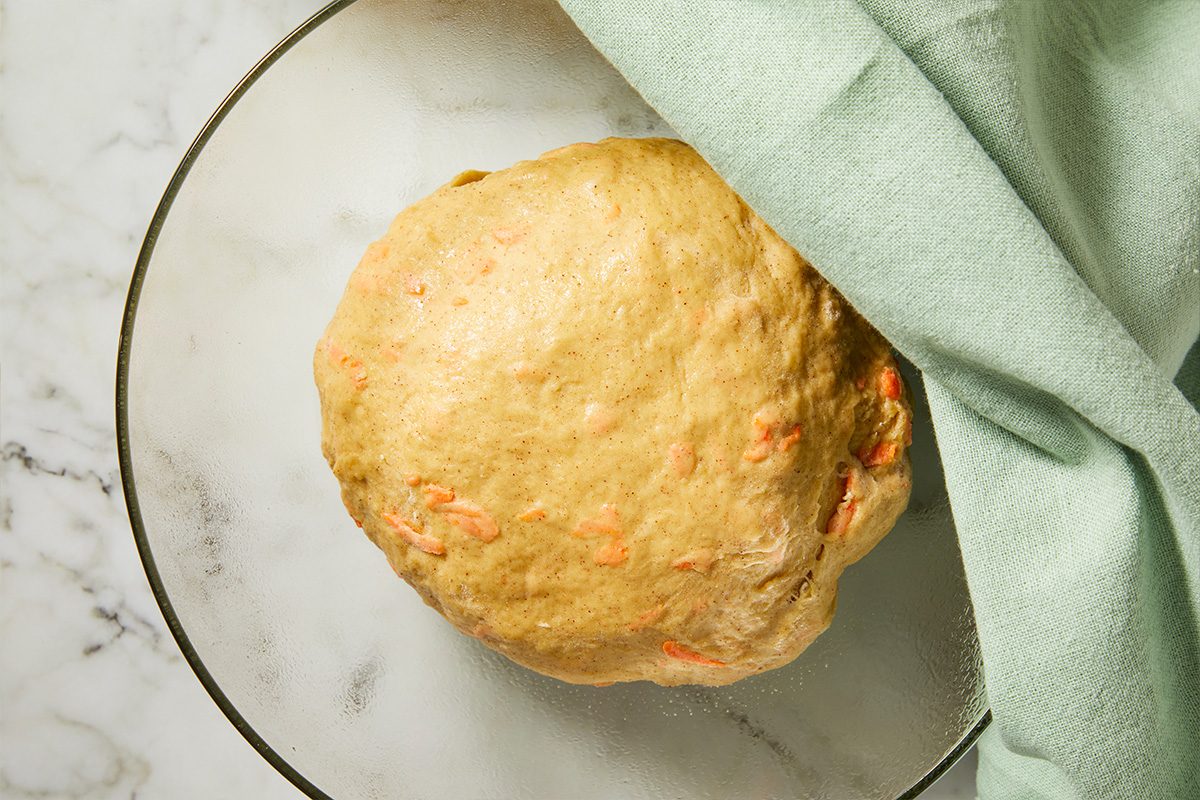 Overhead view of carrot cake cinnamon roll dough formed into a smooth ball and resting in a glass bowl before rising.