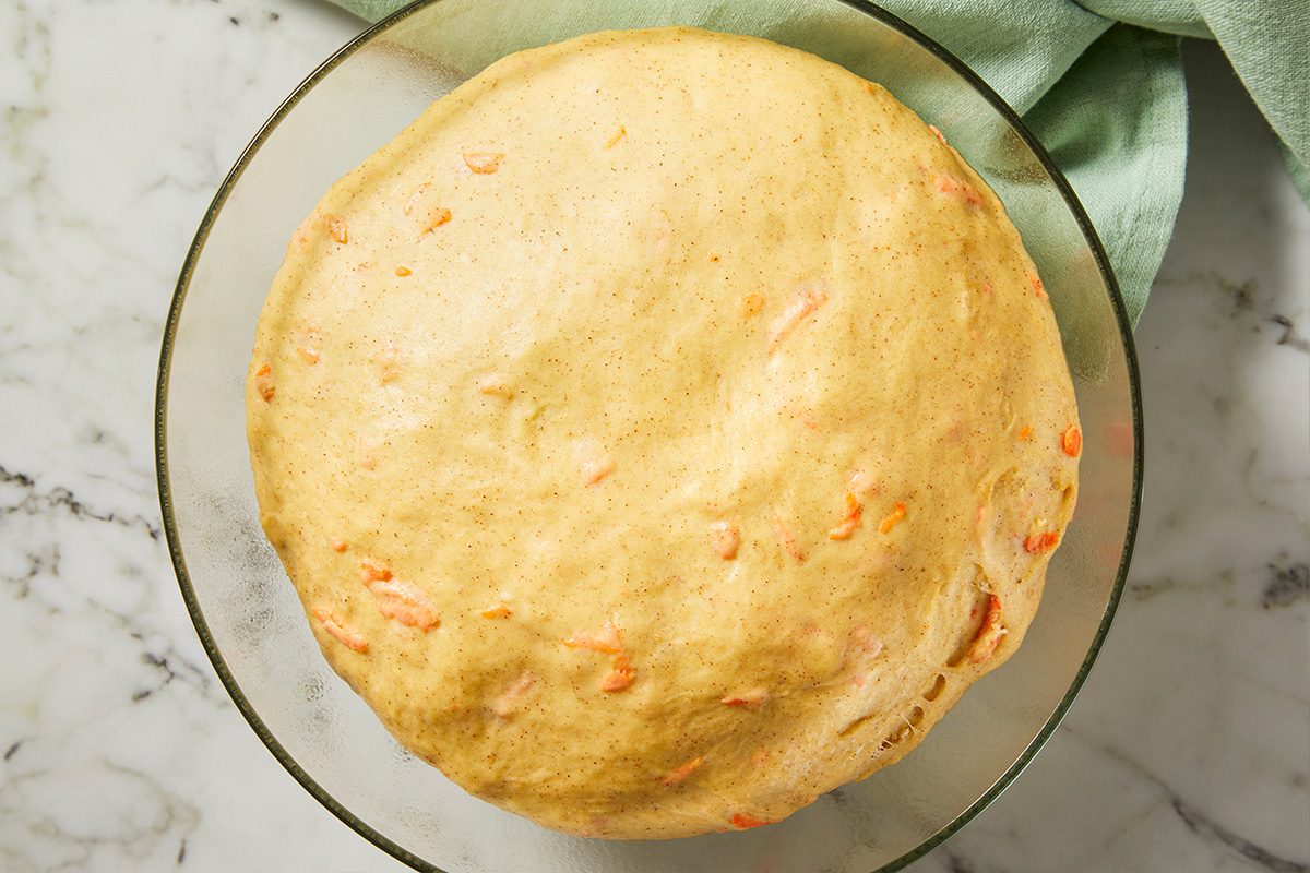 Overhead view of risen carrot cake cinnamon roll dough in a glass bowl, showing an expanded, airy texture after proofing.