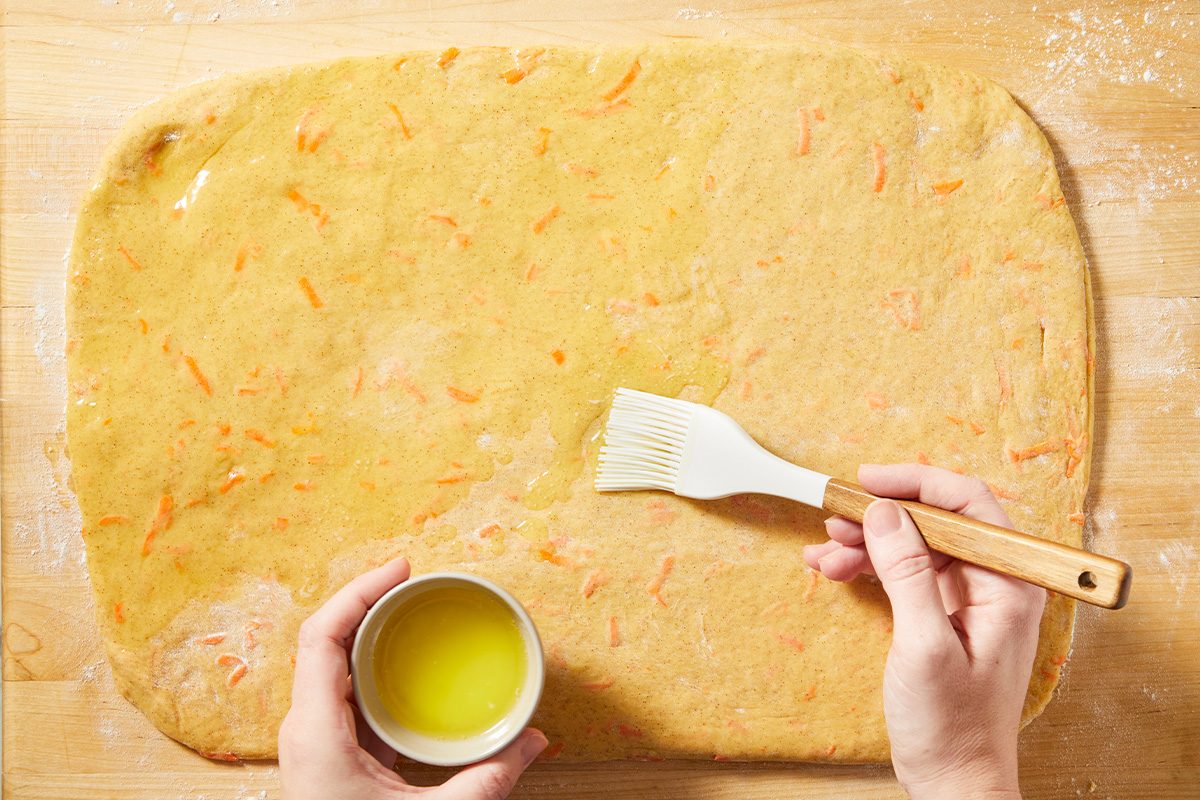 Overhead view of rolled-out carrot cake dough being brushed with melted butter before adding cinnamon sugar filling.
