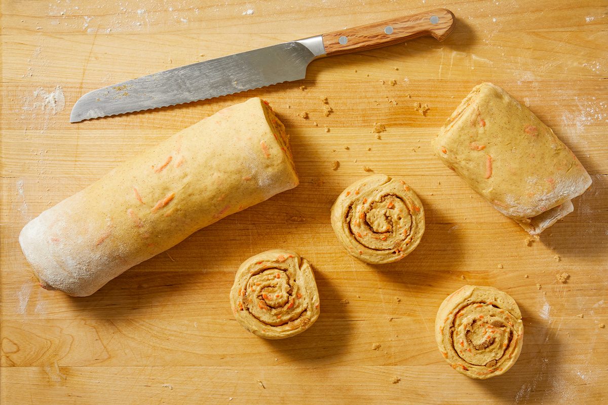 Overhead view of carrot cake dough rolled into a log and sliced into individual cinnamon rolls on a wooden surface.
