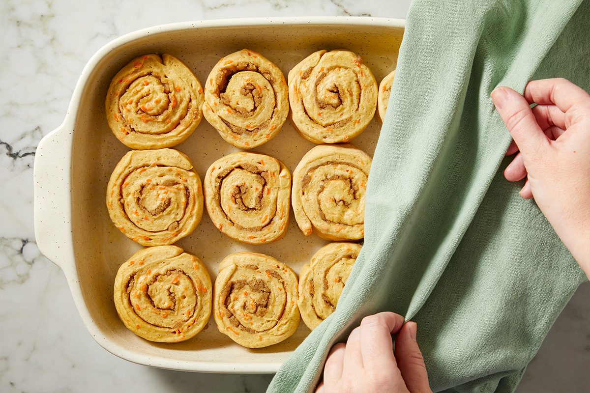 Overhead view of unbaked carrot cake cinnamon rolls arranged in a baking dish and covered with a cloth to rise.