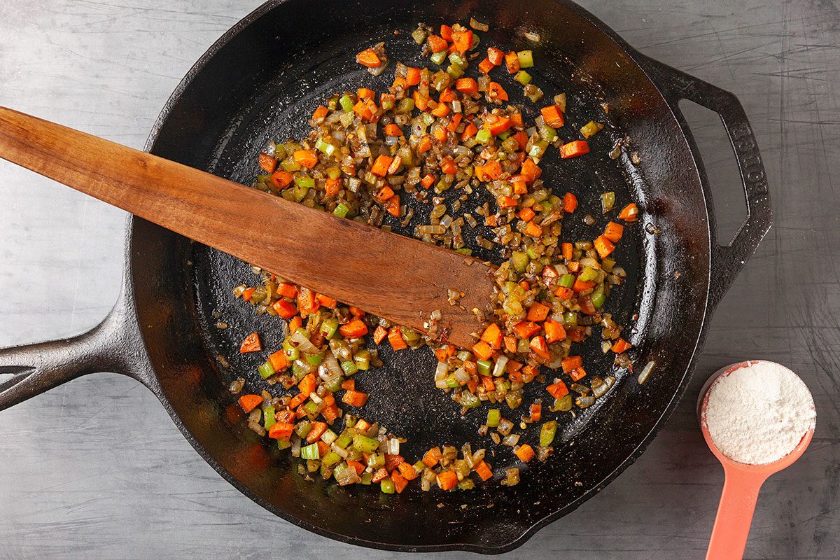 Overhead horizontal image of chopped onion, celery, and carrot sautéing in butter in a cast iron skillet, stirred with a wooden spoon. This image captures the aromatic vegetable base cooking down to build flavor for the pot pie filling.