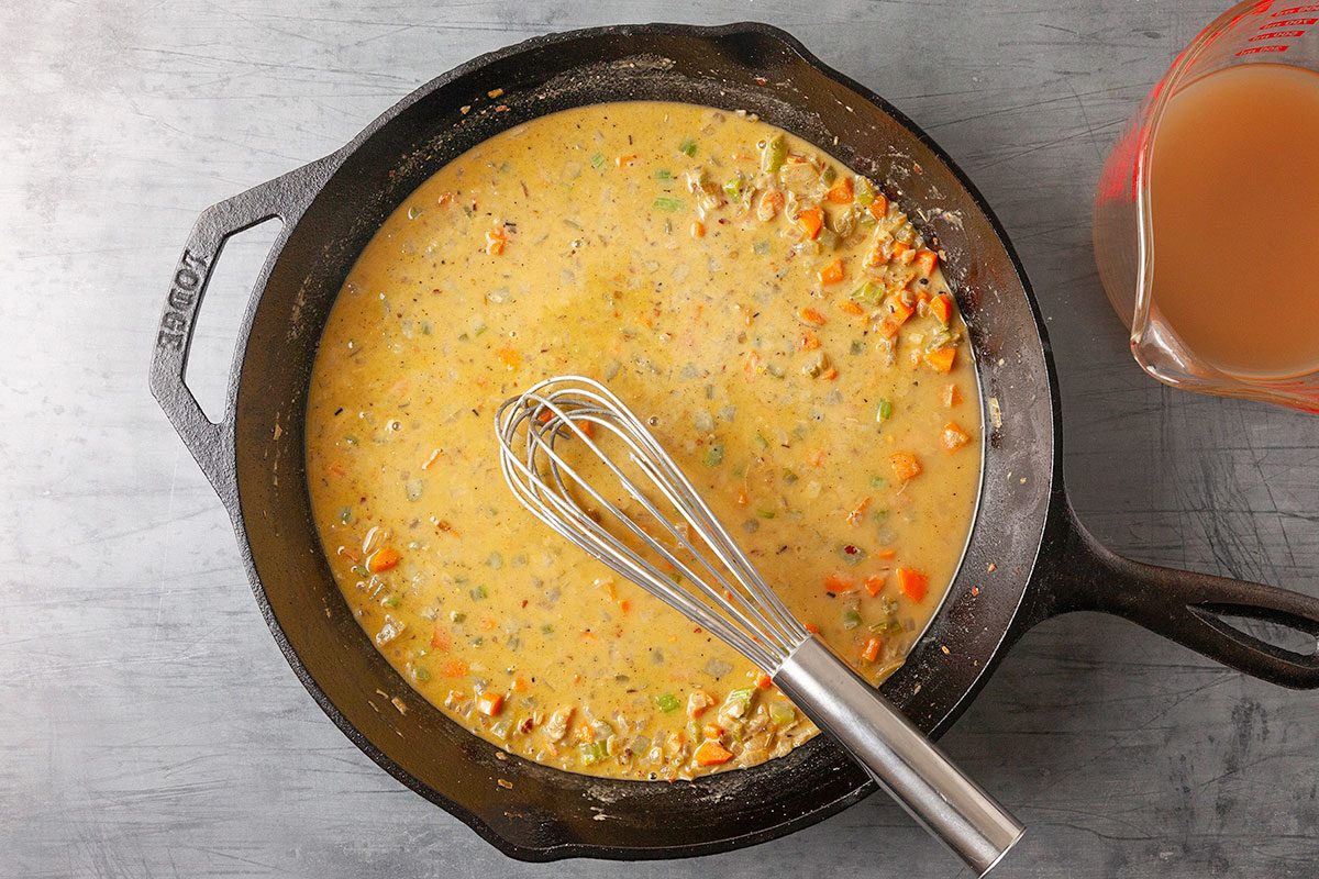 Overhead horizontal image of a creamy pot pie sauce being whisked in a cast iron skillet. Flour, chicken stock, and cream are combined to form a smooth, rich gravy that will bind the filling ingredients.
