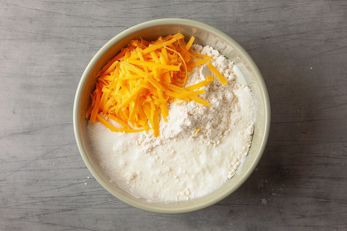 Overhead horizontal image of Cheddar Bay biscuit dough ingredients in a mixing bowl, featuring biscuit mix, shredded sharp cheddar cheese, and dairy components before mixing.