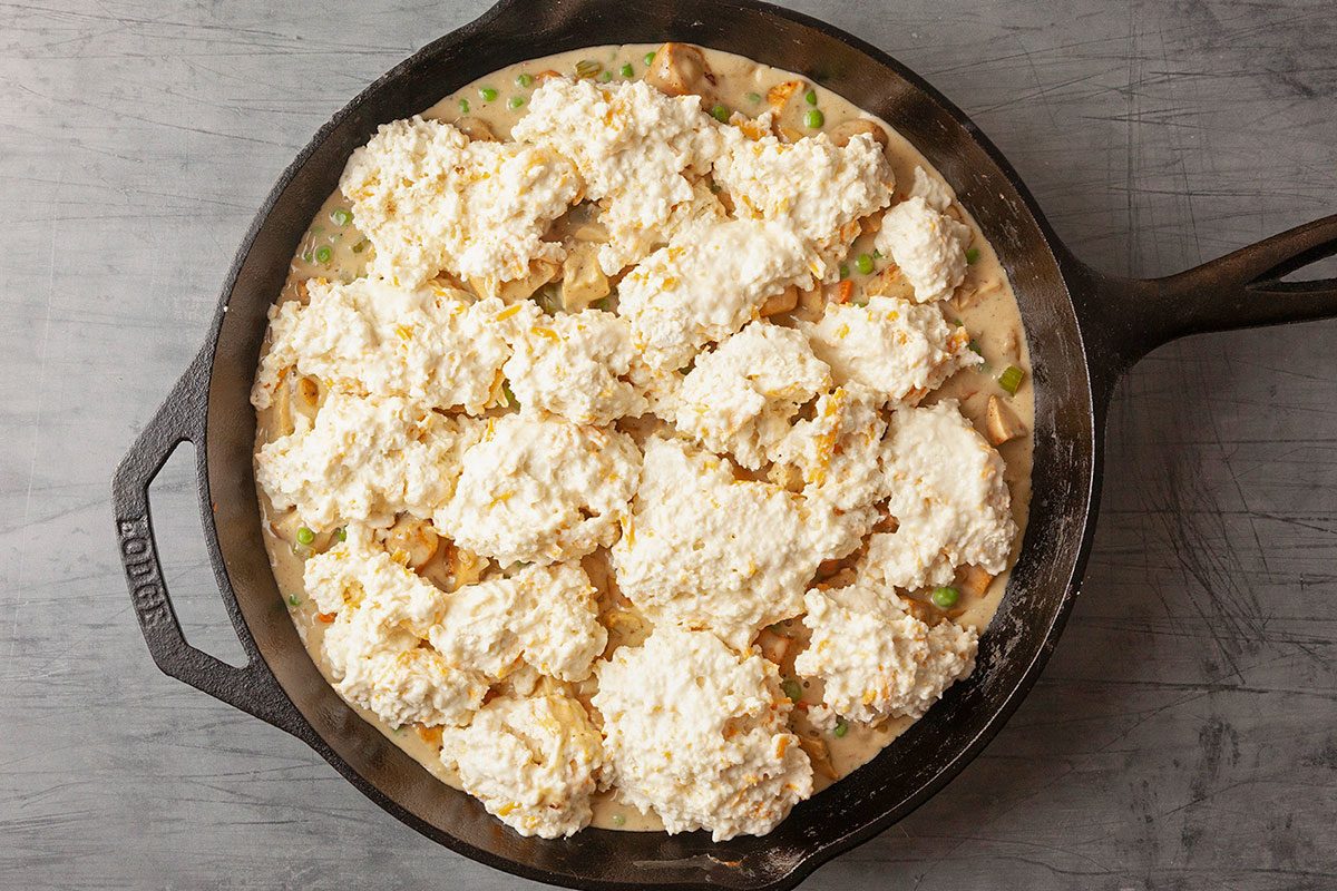 Overhead horizontal image of spooned biscuit dough distributed over the chicken pot pie filling in a cast iron skillet, showing uneven rustic dollops before baking.