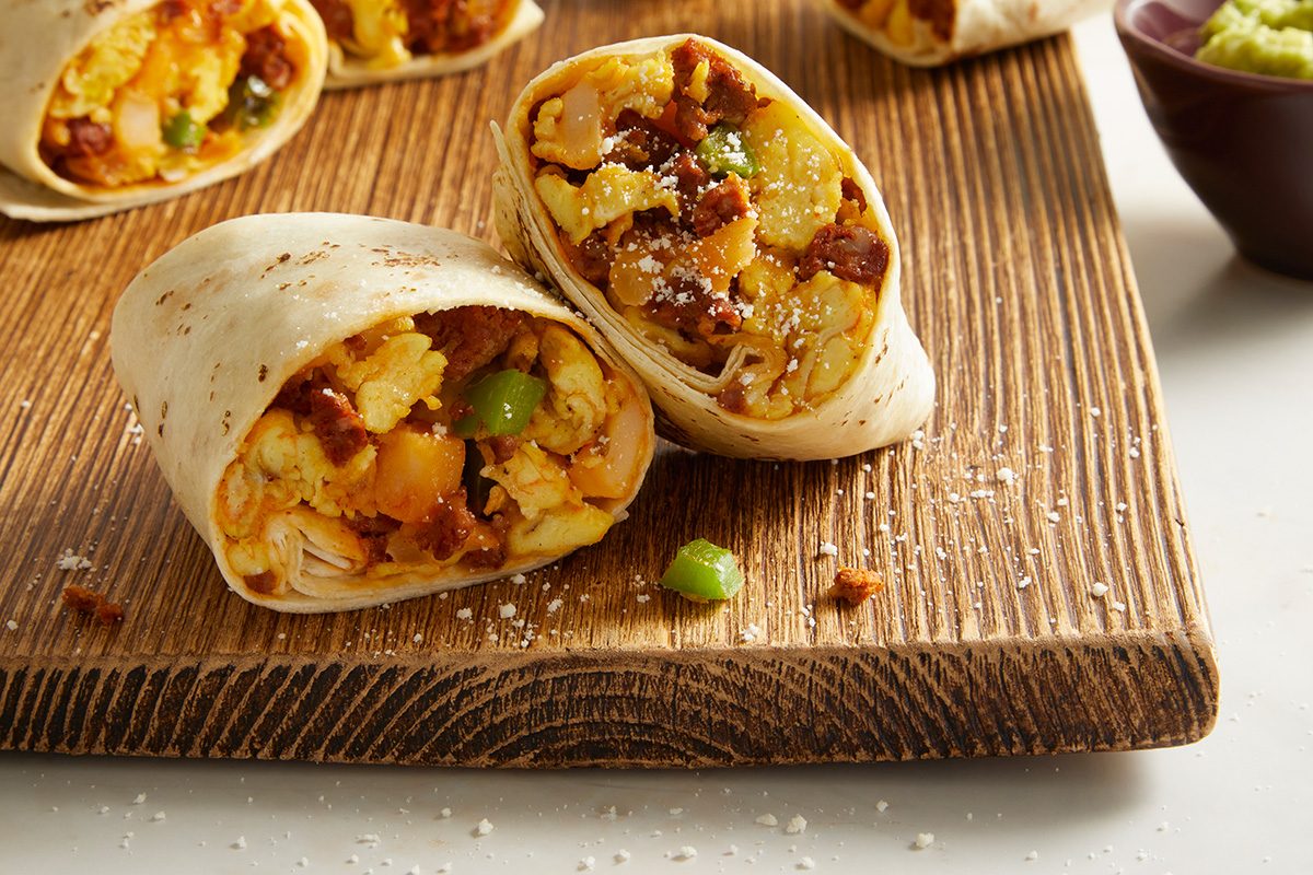 A close-up of a sliced breakfast burrito filled with scrambled eggs, sausage, cheese, and green peppers on a wooden board, with more burritos and a bowl of guacamole in the background.
