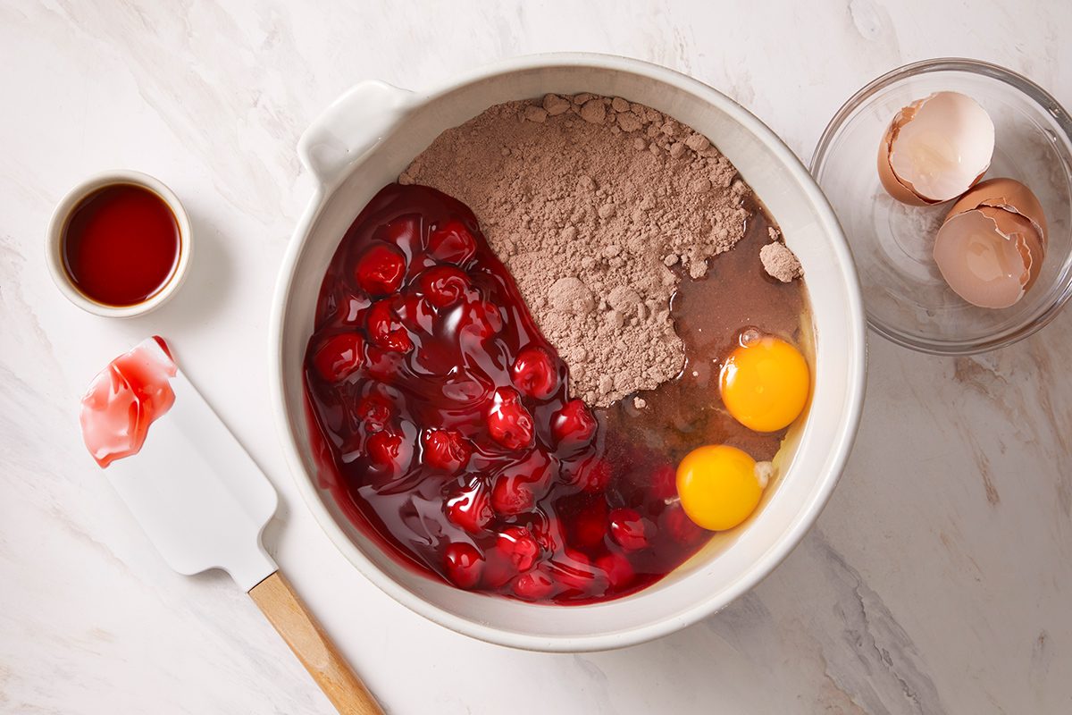 A mixing bowl contains chocolate cake mix, cherry pie filling, and two eggs. Nearby are a spatula with some red filling, a small cup of vanilla extract, and cracked eggshells on a light countertop.