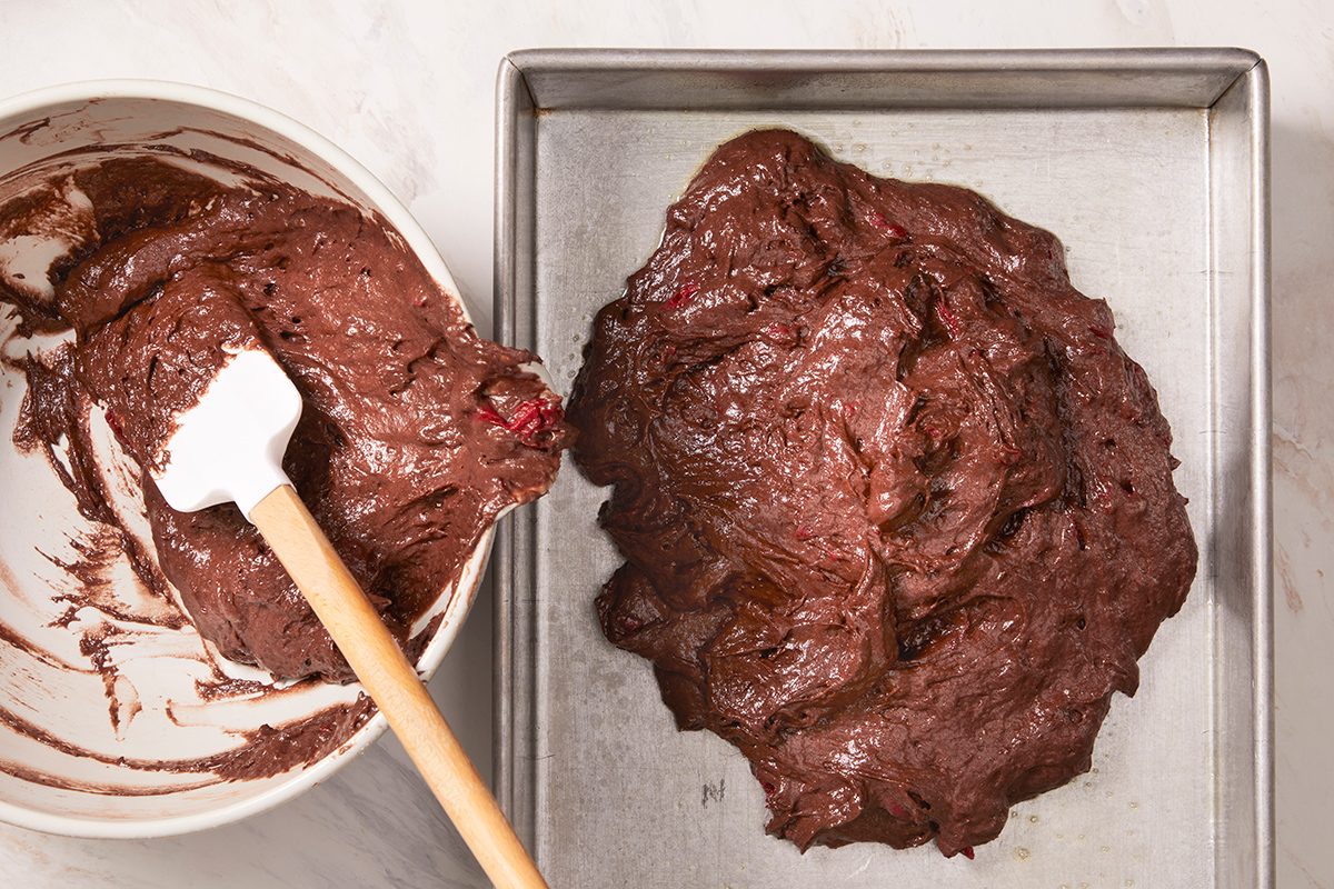 A metal baking pan with thick chocolate brownie batter inside sits next to a mixing bowl with more batter and a white spatula resting on the rim.