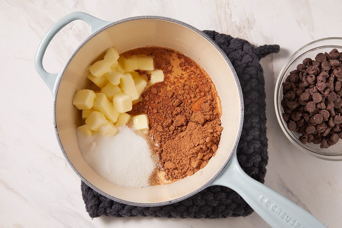 A light blue pot containing cubes of butter, white sugar, and cocoa powder sits on a dark mat, with a bowl of chocolate chips next to it on a white surface.