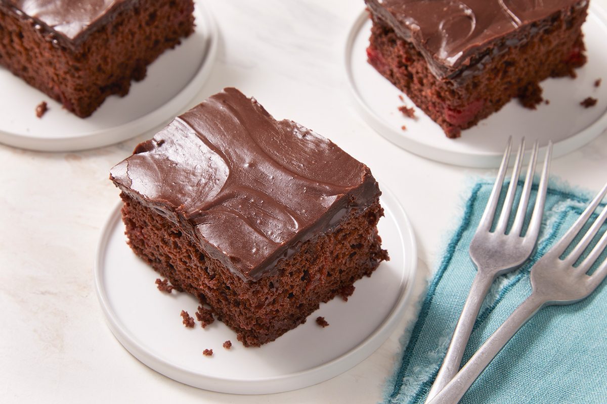 Three pieces of chocolate cake with smooth chocolate frosting are served on small white plates. Two forks rest on a blue napkin beside the plates, all set on a light-colored surface.