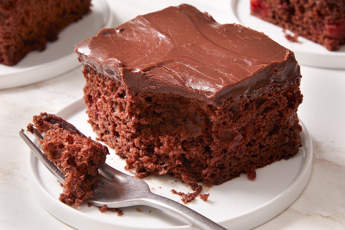 A square slice of chocolate cake with thick chocolate frosting sits on a white plate, with a fork holding a bite-sized piece in front of it. More pieces of cake are visible in the background.