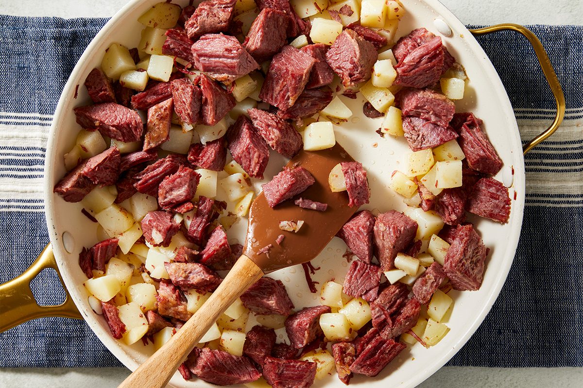 An overhead, horizontal step-by-step image showing chopped cooked corned beef sautéed with refrigerated diced potatoes and onion in a skillet. This mixture forms the filling for Spicy Corned Beef Tacos.