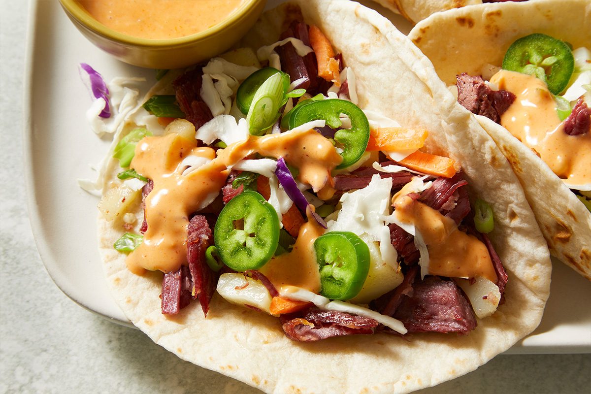 An overhead, horizontal close-up image of a Spicy Corned Beef Taco filled with corned beef and potatoes, topped with coleslaw, sliced jalapeños, and spicy Thousand Island dressing.