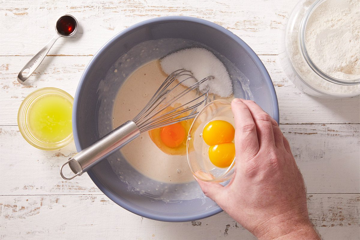 An overhead, horizontal, step-by-step image showing eggs being added to a mixing bowl containing yeast dough ingredients. Warm milk, sugar, flour, and yeast are visible, illustrating an early stage in preparing dough for Crème Brûlée Doughnuts.