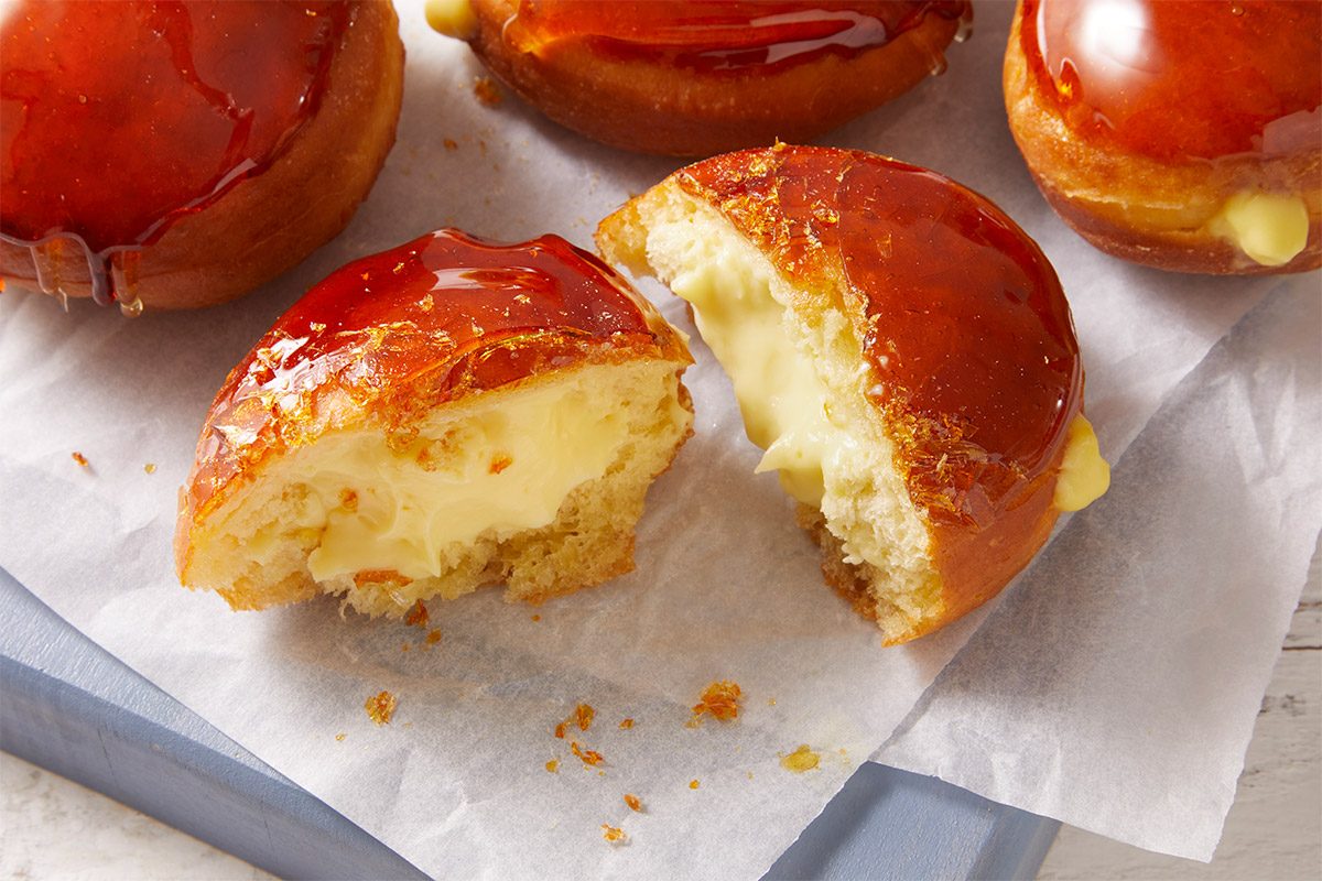 An overhead, horizontal close-up image of a Crème Brûlée Doughnut cut open to reveal creamy pastry cream inside, topped with crisp caramelized sugar.