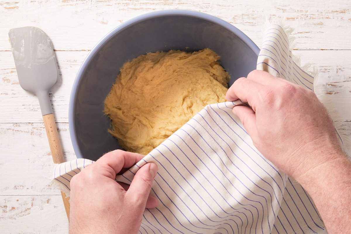 An overhead, horizontal image showing soft dough being covered with a kitchen towel in a mixing bowl as it rests and rises. This step demonstrates yeast activation during the preparation of Crème Brûlée Doughnuts.