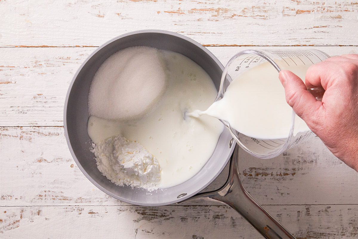A horizontal overhead image showing milk being poured into a saucepan to heat for pastry cream. This step captures preparation of the custard filling used in Crème Brûlée Doughnuts.