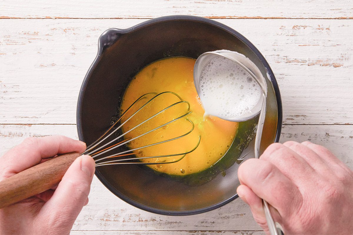 An overhead, horizontal step-by-step image showing egg yolks being whisked with sugar in a saucepan. This mixture forms the base of the pastry cream filling for Crème Brûlée Doughnuts.