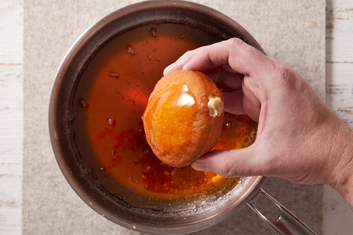A horizontal overhead image showing a doughnut being dipped into caramel made from sugar and water. This step creates the signature brûléed topping for Crème Brûlée Doughnuts.