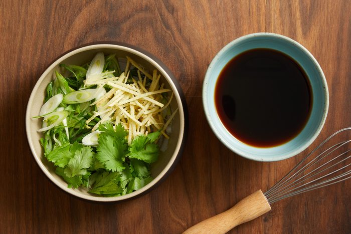sliced green onions, peeled and julienned ginger and cilantro sprigs in a bowl next to soy sauce mixture