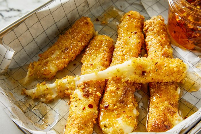 Closeup shot of Hot Honey Fried Mozzarella; golden, crispy cheese sticks glistening with chili-flecked hot honey; one stick pulled apart to reveal a long, stretchy melted cheese pull; served on a parchment-lined tray with a jar of hot honey visible in the corner;