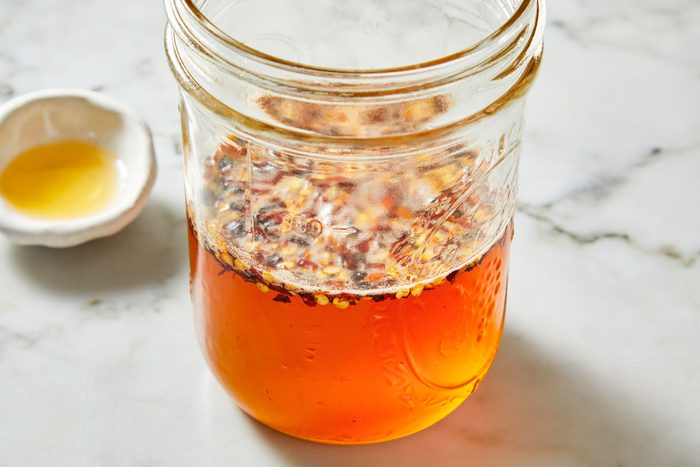 Closeup shot of a glass jar filled with homemade chili oil; vibrant red chili flakes suspended in bright orange oil; set on a white marble surface with a small dish of yellow oil blurred in the background;