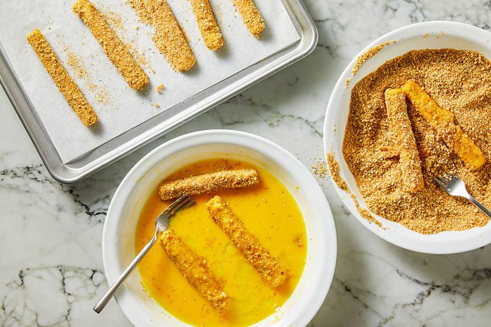 Overhead shot of breading cheese sticks; one bowl holds beaten eggs, another is filled with seasoned breadcrumbs, and a parchment-lined tray shows coated sticks; forks rest in the bowls as the cheese sticks are dipped and coated on a marble surface;