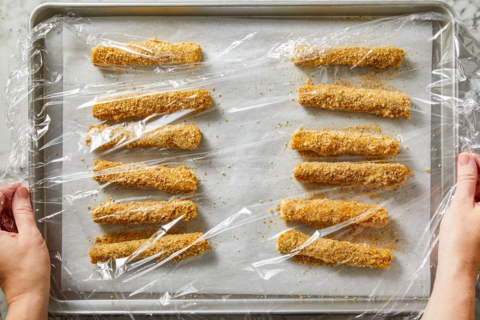 Overhead shot of breaded cheese sticks arranged in neat rows on a parchment-lined baking sheet; hands pull plastic wrap over the tray to cover the coated sticks;
