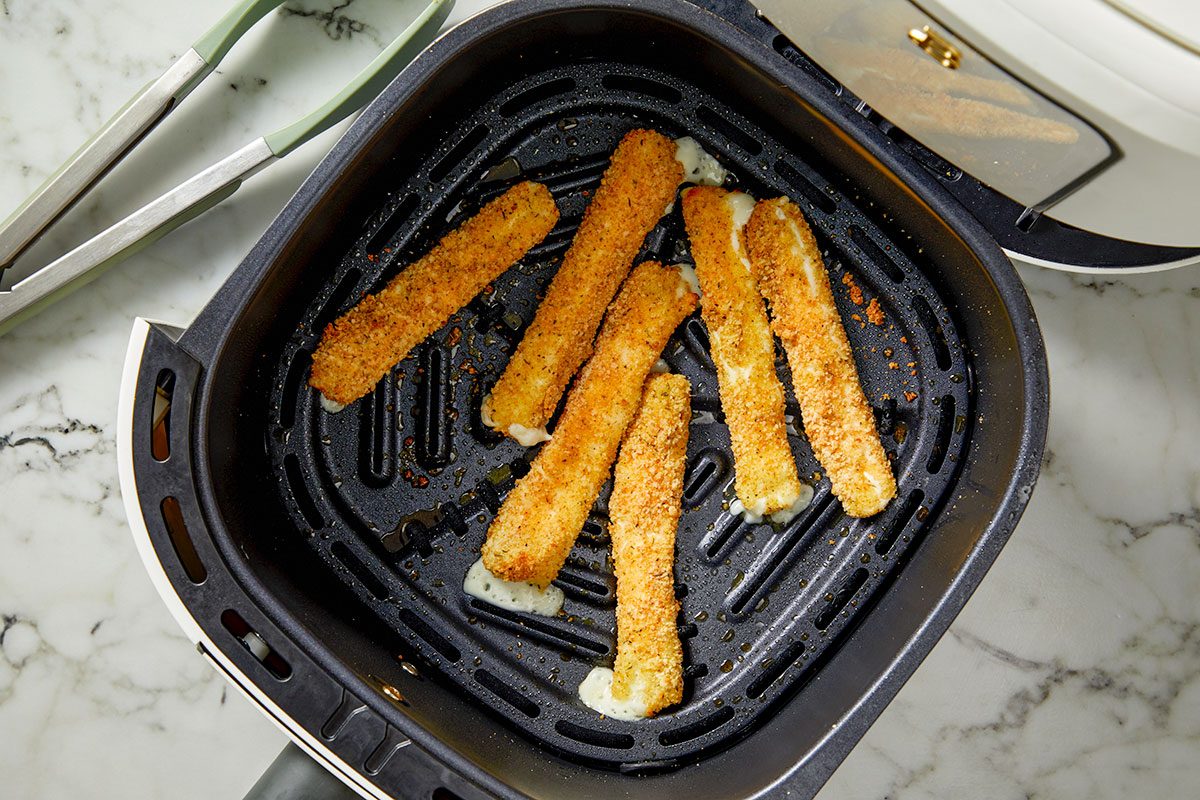 Overhead shot of air-fried cheese sticks in an air fryer basket; golden and crisp with small spots of melted cheese oozing out; tongs rest on the marble countertop beside the fryer;