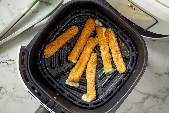 Overhead shot of air-fried cheese sticks in an air fryer basket; golden and crisp with small spots of melted cheese oozing out; tongs rest on the marble countertop beside the fryer;