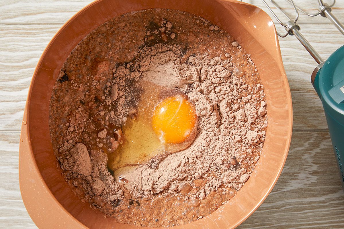 Overhead view of dry cake mix ingredients in a mixing bowl with a cracked egg added at the center, marking the first step in preparing the Irish cream poke cake batter on a light wood surface.