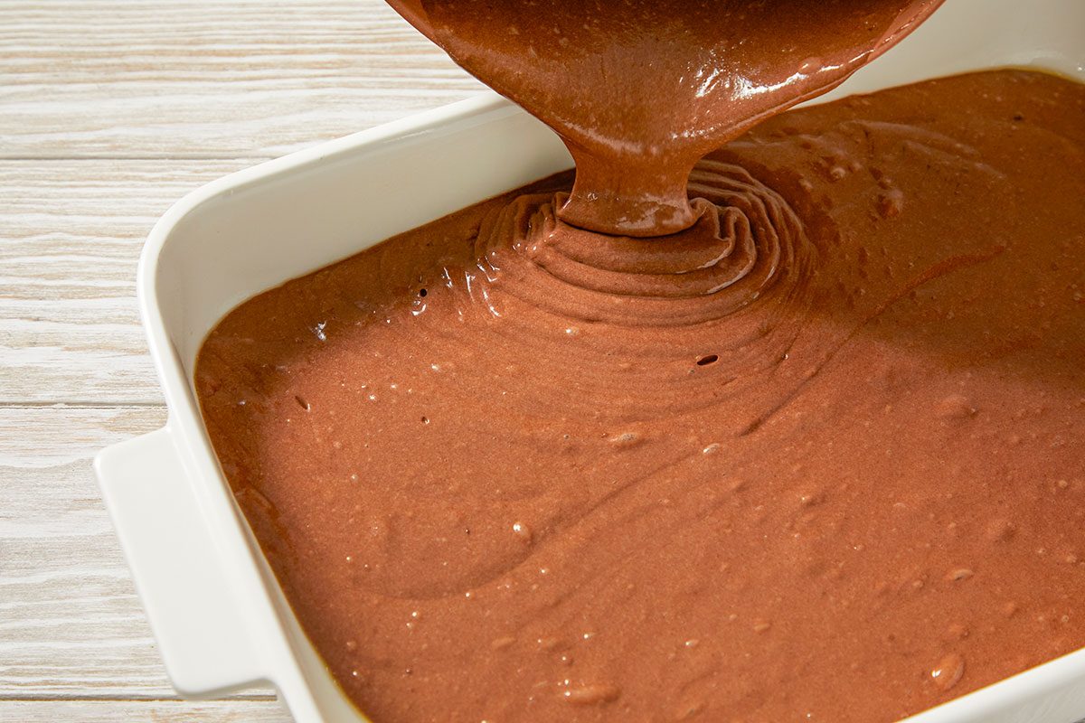 Top-down image of smooth chocolate cake batter being poured into a rectangular baking dish, showing an early preparation step for the Irish cream poke cake.
